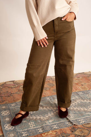 A person wearing olive brown wide-leg pants with a cream ribbed sweater and burgundy flats, standing on a patterned rug against a plain beige wall.