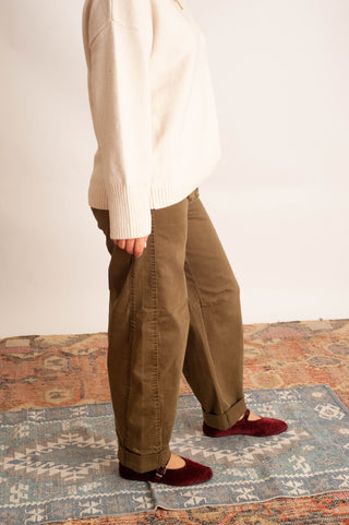 A person shown from the chest down wearing loose olive-brown pants, a cream sweater, and burgundy flats, standing on a patterned rug against a plain light wall.
