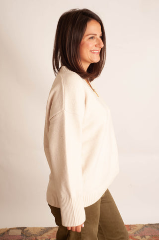 A woman with shoulder-length dark brown hair is shown in profile, wearing an oversized cream-colored knit sweater and olive green pants against a plain off-white background.