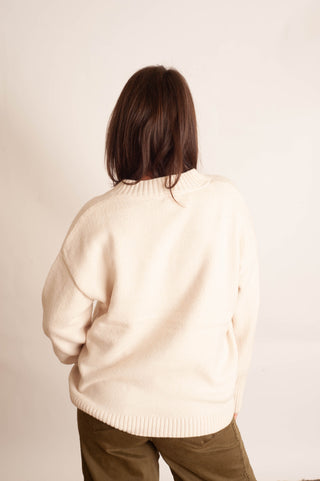 A person viewed from behind wearing a cream-colored knit sweater and olive green pants against a plain off-white background.