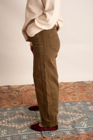 A person shown from the waist down wearing olive brown straight-leg corduroy pants, a cream knit sweater, and burgundy flats, standing on a patterned rug against a plain off-white wall.