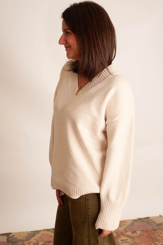 A woman wearing a cream-colored knit sweater with a V-neck and ribbed cuffs, paired with olive green pants, standing against a plain off-white wall.
