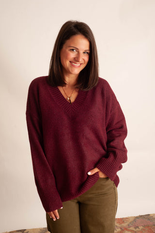 A woman wearing a maroon V-neck knit sweater and olive green pants, standing against a plain off-white wall.