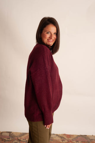 A woman wearing an oversized burgundy knit sweater and olive green pants, standing against a plain off-white wall.