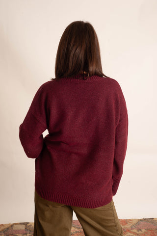 A person viewed from behind wearing an oversized maroon knit sweater and olive green pants against a plain off-white background.