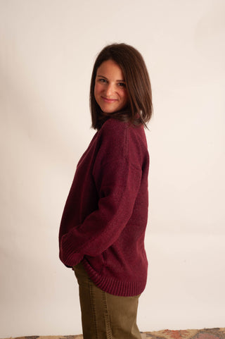 A woman with shoulder-length brown hair is shown from the back and side, wearing a maroon knit sweater and olive green pants against a plain off-white background.