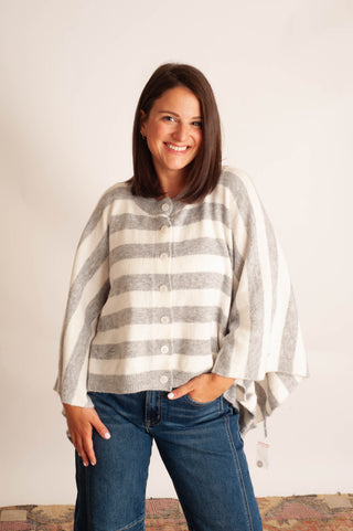 A woman wearing a gray and white striped knit poncho cardigan with blue jeans, standing against a plain off-white wall.