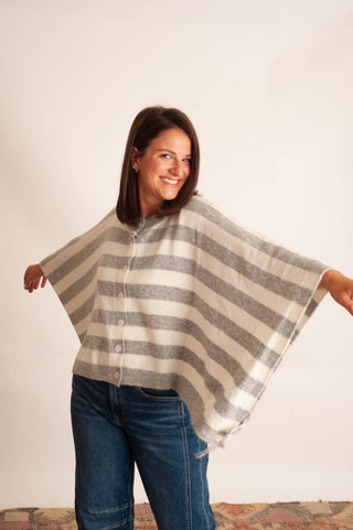 A woman modeling a cream and gray striped button-up poncho with wide sleeves, paired with blue jeans, against a plain off-white background.