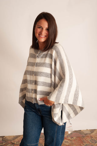 A woman wearing a cream and gray striped button-up poncho cardigan with blue jeans, standing against a plain off-white wall.