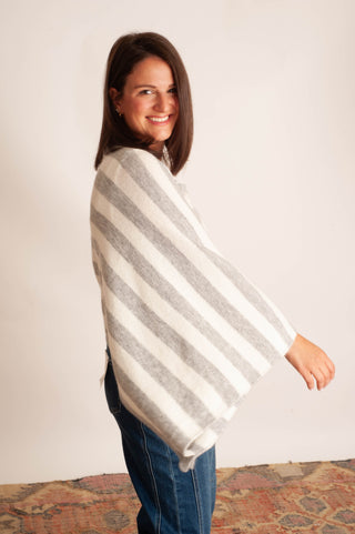 A woman wearing a cream and gray striped knit poncho sweater, paired with blue jeans, standing against a plain off-white wall.