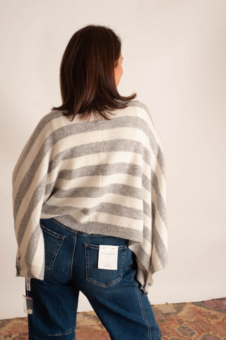 A woman viewed from behind wearing a cream and gray striped knit poncho sweater and blue jeans, standing against a plain off-white wall.