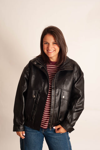 A woman wearing a black leather jacket over a burgundy and white striped shirt, paired with blue jeans, standing against a plain off-white background.