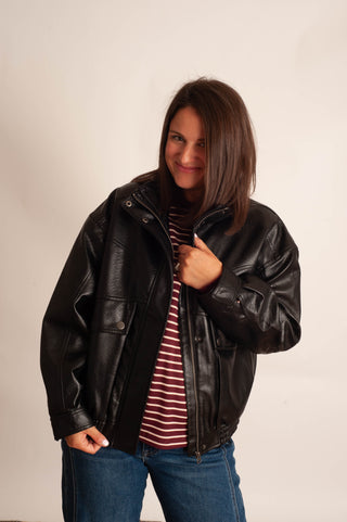 A woman wearing a black faux-leather moto jacket over a red and white striped t-shirt and blue jeans, posing against a plain off-white background.