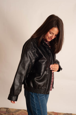 A woman wearing a black faux-leather moto jacket over a red and white striped shirt and blue jeans, standing against a plain off-white wall.