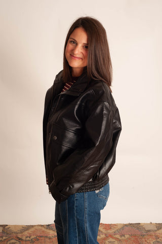 A woman with long brown hair wearing a black leather jacket, blue jeans, and a striped shirt, standing against a plain off-white wall.