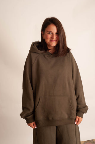 A woman wearing an oversized olive brown pullover hoodie with a kangaroo pocket, paired with matching wide-leg sweatpants, standing against a plain off-white background.