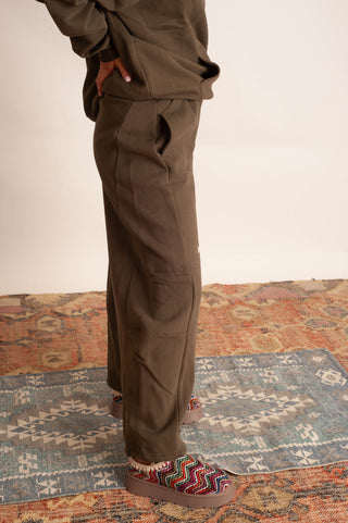 A person wearing loose brown pants and a matching brown top, standing on a patterned rug with colorful slip-on shoes. The photo is cropped from the chest down and focuses on the pants.