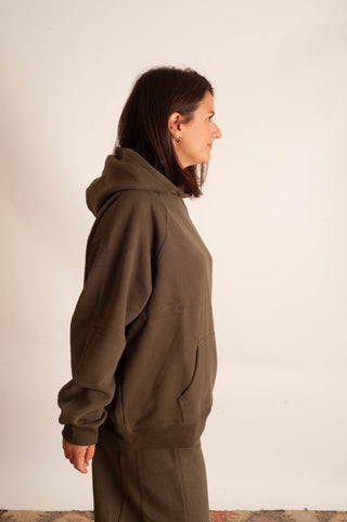 A woman shown in profile wearing a dark brown pullover hoodie and matching pants against a plain off-white wall.