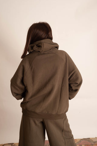 A person shown from behind wearing a loose brown hooded sweatshirt and matching brown pants against a plain off-white wall.
