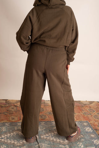 A person shown from the back wearing a matching dark olive brown cropped hoodie and wide-leg sweatpants, standing on a patterned rug against a plain wall.
