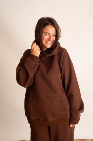 A woman wearing a brown pullover hoodie and matching brown pants, smiling and posing against a plain off-white studio background.