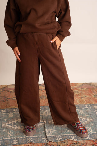 A person wearing a matching brown sweatshirt and wide-leg sweatpants, standing on a patterned rug with colorful slip-on shoes. The photo is cropped from the chest down against a plain light background.