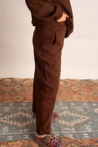 A person wearing brown drawstring sweatpants and a matching brown top, standing on a patterned rug against a plain light wall.