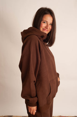 A woman wearing an oversized brown hoodie and matching brown pants, photographed against a plain off-white studio background.