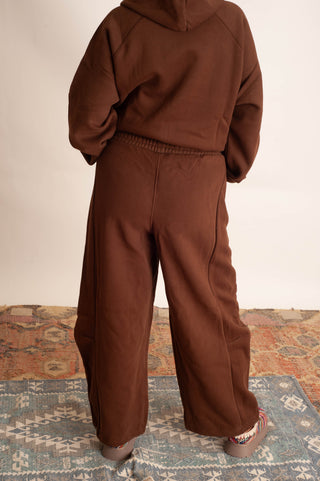 A person viewed from behind wearing a matching brown hoodie and wide-leg sweatpants set, standing on a patterned rug against a plain light wall.