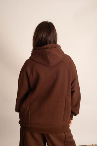 A person viewed from behind wearing a dark brown pullover hoodie and matching brown pants against a plain off-white wall.