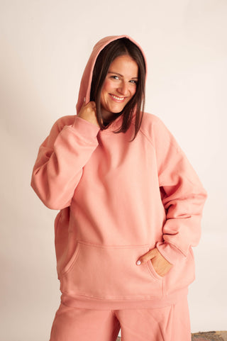 A woman wearing a matching dusty pink loungewear set, featuring a pullover hoodie with a front kangaroo pocket and matching sweatpants, posed against a plain off-white background.