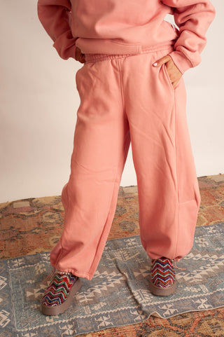 A person wearing a matching dusty pink sweatshirt and wide-leg jogger pants, standing on a patterned rug. The outfit is shown from the chest down, with hands in the pockets of the pants.