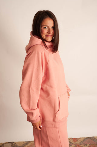A woman wearing a matching dusty pink loungewear set, featuring a pullover hoodie and coordinating pants, standing against a plain off-white wall.
