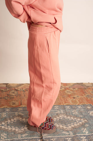 A person wearing dusty pink sweatpants and a matching sweatshirt, standing on a patterned rug against a plain light wall.