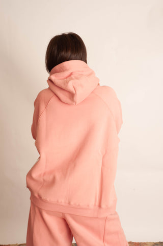A person shown from behind wearing a light peach-pink hooded sweatshirt and matching sweatpants against a plain off-white background.