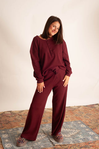 A woman modeling a matching burgundy lounge set consisting of a long-sleeve drawstring top and wide-leg pants, standing on a patterned rug against a plain off-white wall.