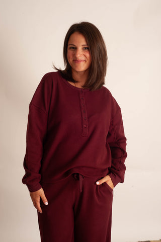 A woman wearing a matching burgundy lounge set consisting of a long-sleeve henley sweatshirt and drawstring sweatpants, standing against a plain off-white background.