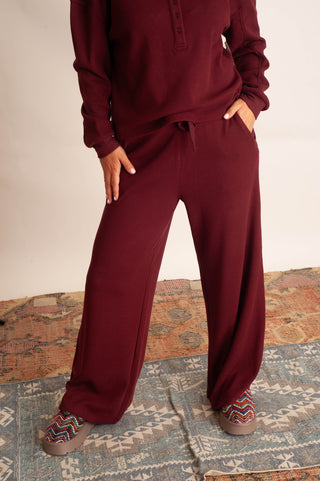 A person wearing a matching burgundy lounge set with a long-sleeve henley top and wide-leg drawstring pants, standing on a patterned rug against a plain light wall.