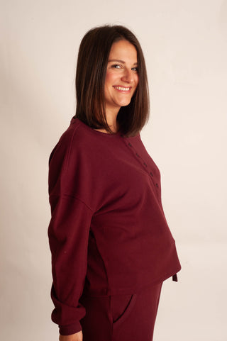 A woman with shoulder-length brown hair is shown from the side, wearing a burgundy long-sleeve cropped henley sweatshirt and matching sweatpants against a plain off-white studio background.