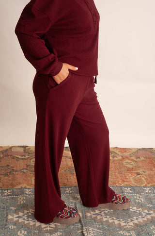 A person wearing a matching burgundy lounge set with a long-sleeve top and wide-leg pants, standing on a patterned rug against a plain beige wall.