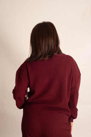 A woman viewed from behind wearing a matching burgundy sweatshirt and sweatpants set against a plain off-white studio background.