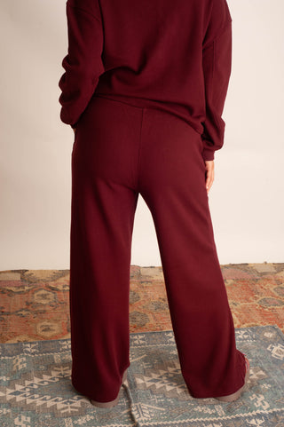 A person shown from the back wearing a matching burgundy sweatshirt and wide-leg sweatpants set, standing on a patterned rug against a plain light wall.