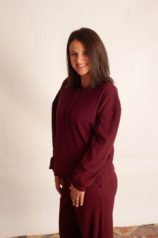 A woman with shoulder-length brown hair stands against a plain off-white wall wearing a matching burgundy lounge set consisting of a long-sleeve top and pants.