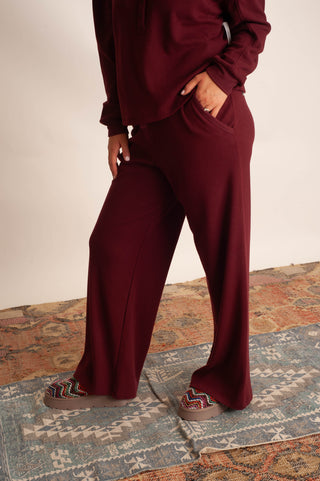 A person wearing a matching burgundy lounge set with wide-leg pants and a long-sleeve top, standing on a patterned rug against a plain light wall.