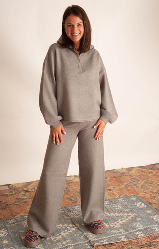 A woman modeling a matching taupe gray lounge set consisting of a long-sleeve half-zip pullover and wide-leg pants, standing on a patterned rug against a plain off-white wall.