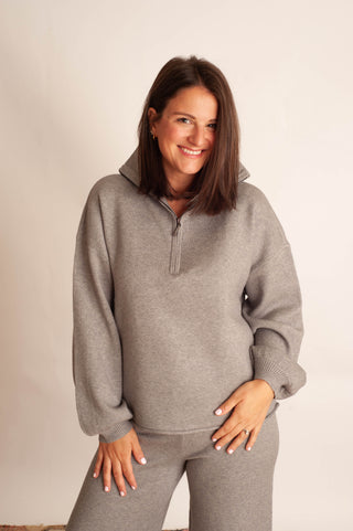 A woman modeling a light gray heathered quarter-zip sweatshirt and matching sweatpants against a plain off-white background.