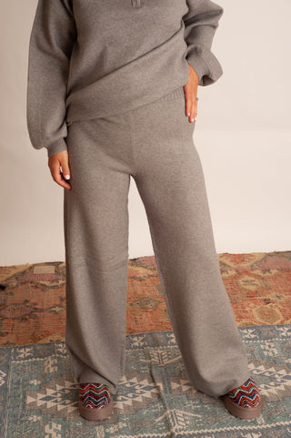 A person wearing a heather gray knit lounge set consisting of a long-sleeve quarter-zip top and matching wide-leg pants, standing on a patterned rug.
