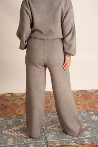 A person shown from the back wearing a matching taupe knit sweater and wide-leg knit pants, standing on a patterned rug against a plain light wall.