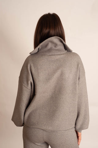 A woman viewed from behind wearing a light gray oversized knit top with a wide folded collar and matching gray bottoms, against a plain light beige studio background.