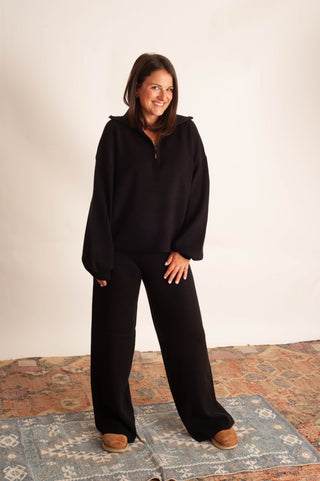 A woman modeling a black half-zip pullover sweatshirt and matching wide-leg pants, standing on a patterned rug against a plain off-white studio background.